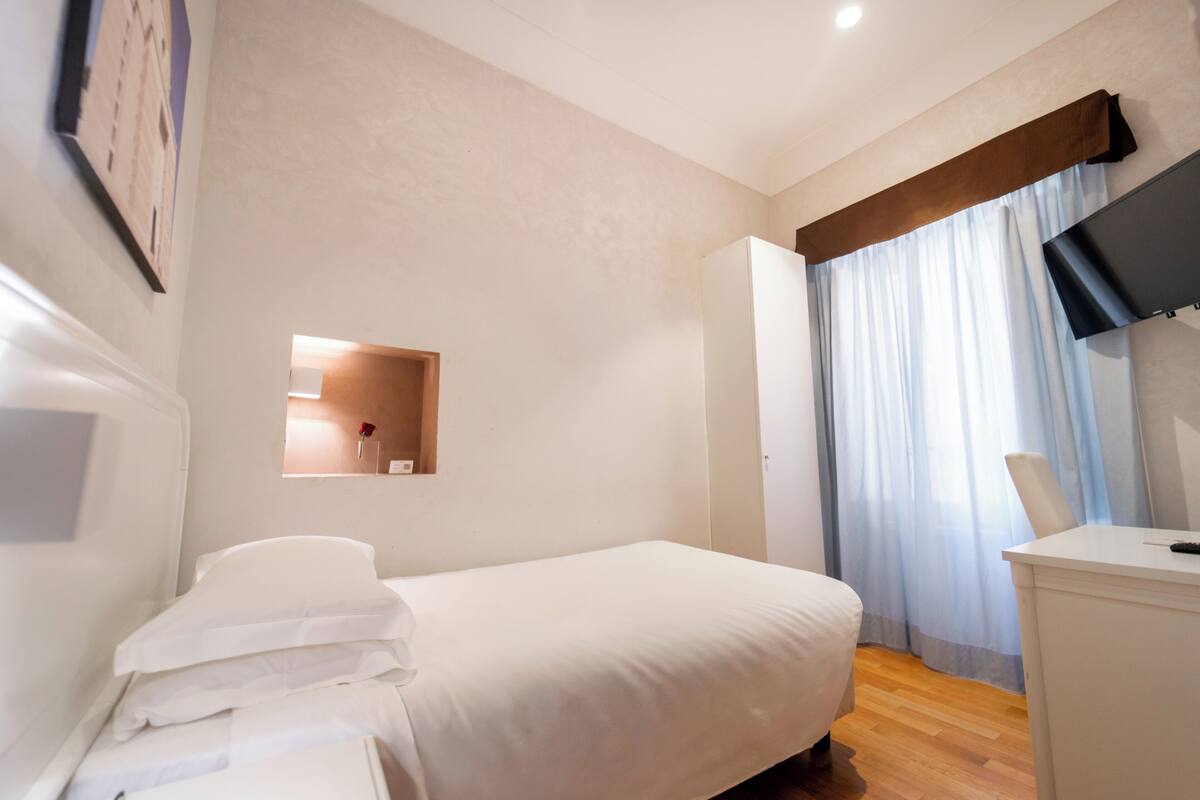 A bedroom with a bed, a desk, a television, and a window with curtains.