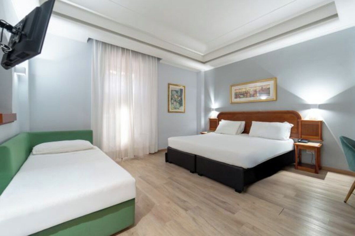 A hotel room with a bed, a sofa, a TV, a window with curtains, and a painting on the wall.