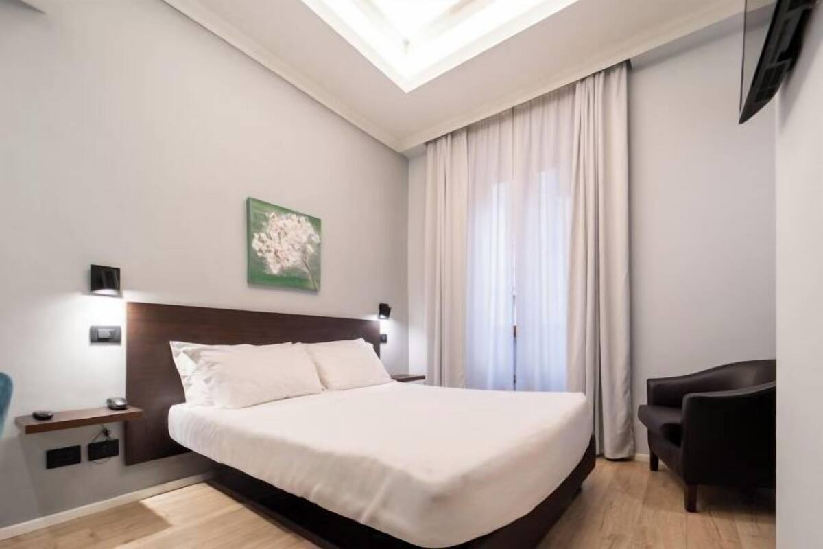A modern hotel room with a large bed, a desk, a chair, a painting on the wall, and a skylight.
