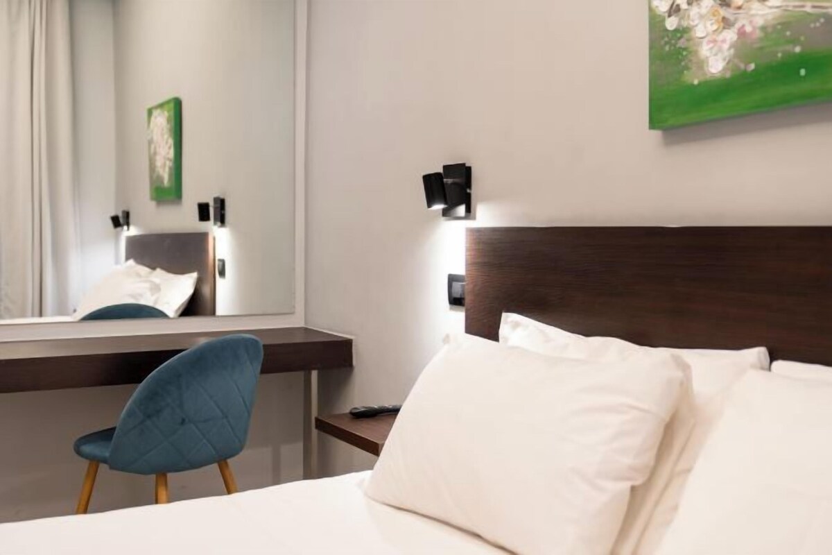 A hotel room with a bed, a desk, a chair, a mirror, and a painting on the wall.