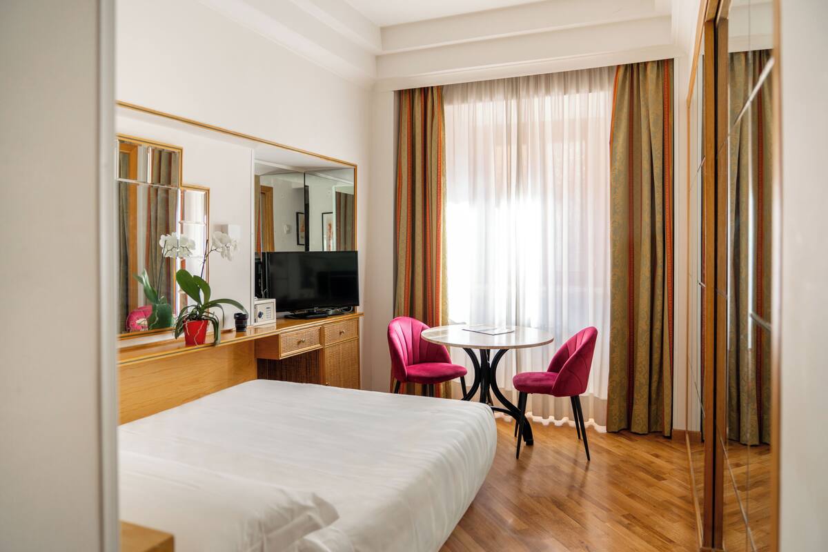 A hotel room with a bed, a desk with a TV, a small table with two chairs, and a large window with curtains.