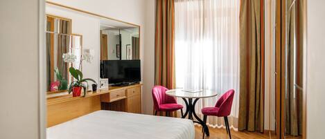 Ett hotellrum med en säng, ett skrivbord med en tv, ett litet bord med två stolar och ett stort fönster med gardiner.