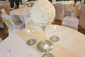 Una mesa redonda con un centro de mesa de cristal decorativo, manteles blancos y pétalos de flores rosadas esparcidos sobre la mesa.