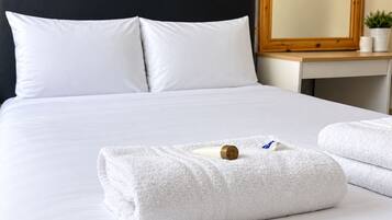 A neatly made bed with white linens and two pillows, a wooden headboard, a bedside table with a plant, and a mirror on the wall.