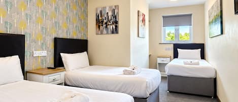 Une chambre d’hôtel avec trois lits, un tableau représentant un paysage urbain, une fenêtre avec vue et un papier peint à motifs.
