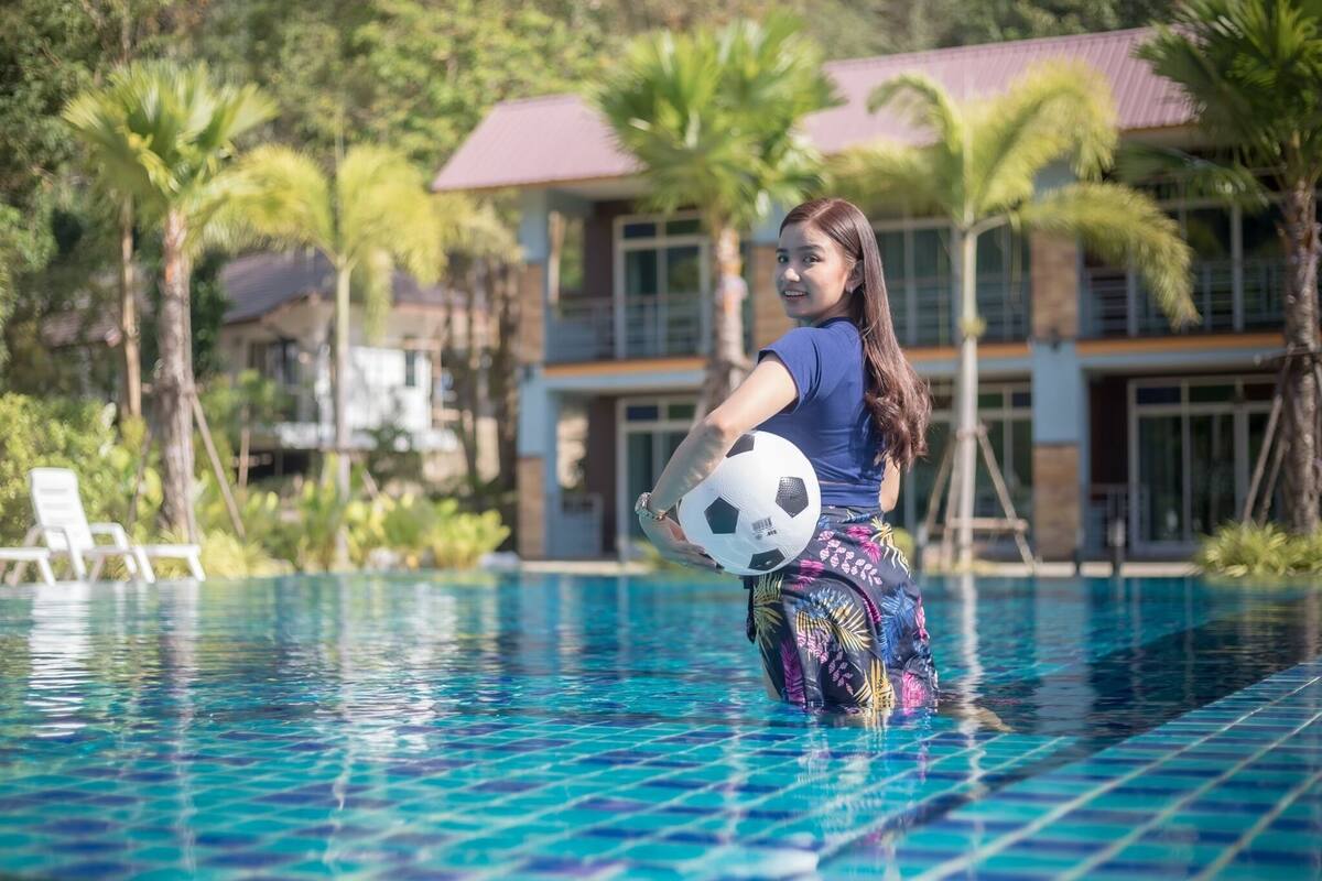 Une personne tenant un ballon de football près d’une piscine, avec des palmiers et des bâtiments en arrière-plan.