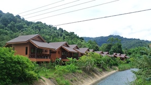 A row of bungalow-style buildings with sloped roofs, situated beside a river, surrounded by lush greenery and hills. - Khaosok Rainforest Resort (Phanom)