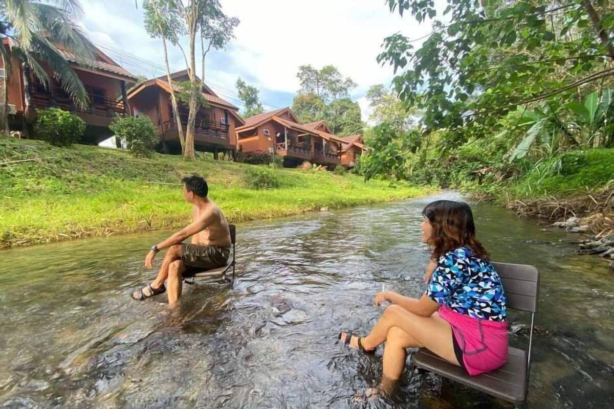 Deux personnes sont assises sur des bancs au bord d’un ruisseau peu profond, entourées d’une végétation luxuriante et de maisons en bois.