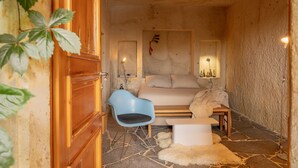 A cozy room with a bed, a blue chair, a small table, and a stone floor. - Serinn House (Urgup)