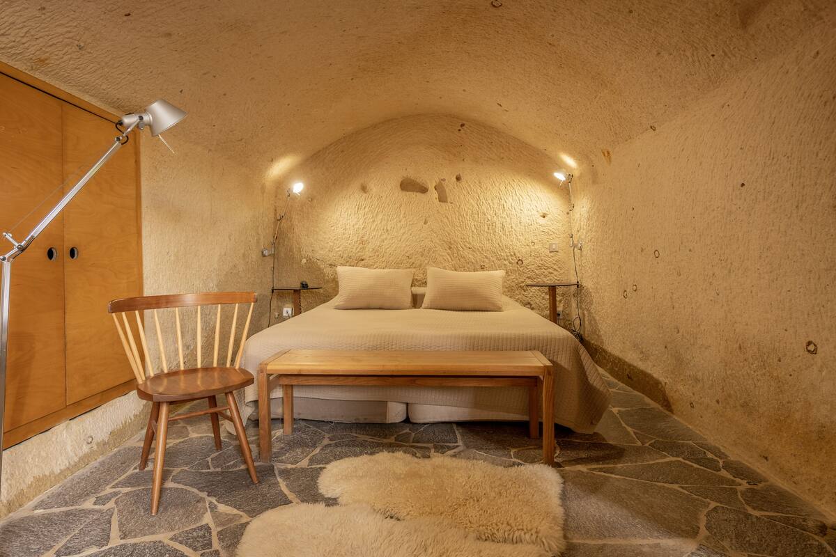 A cozy bedroom with a stone floor, a wooden bed, a chair, a bench, and a lamp.