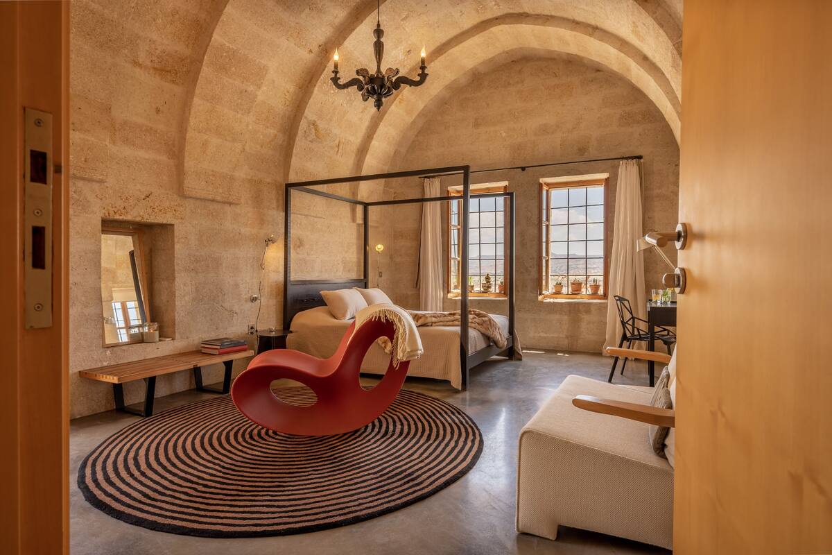 A bedroom with a four-poster bed, a red chair, a round rug, and a view of the outdoors through large windows.