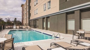 Une piscine entourée de chaises longues et de tables, elle-même bordée d’un bâtiment doté de nombreuses fenêtres et d’un toit plat.