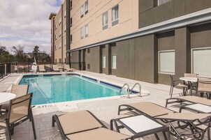 Una piscina con sillas de descanso y mesas, rodeada por un edificio de múltiples ventanas y techo plano.