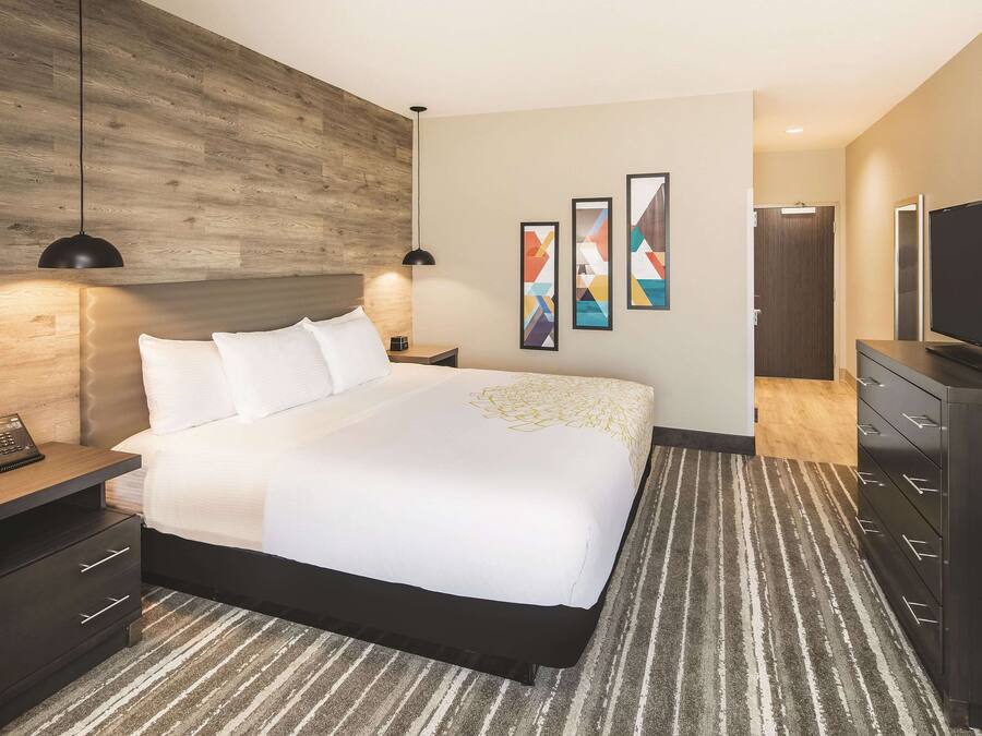 Quarto de hotel com uma cama grande, mesa de cabeceira com telefone, cômoda, televisão e parede decorativa de madeira.