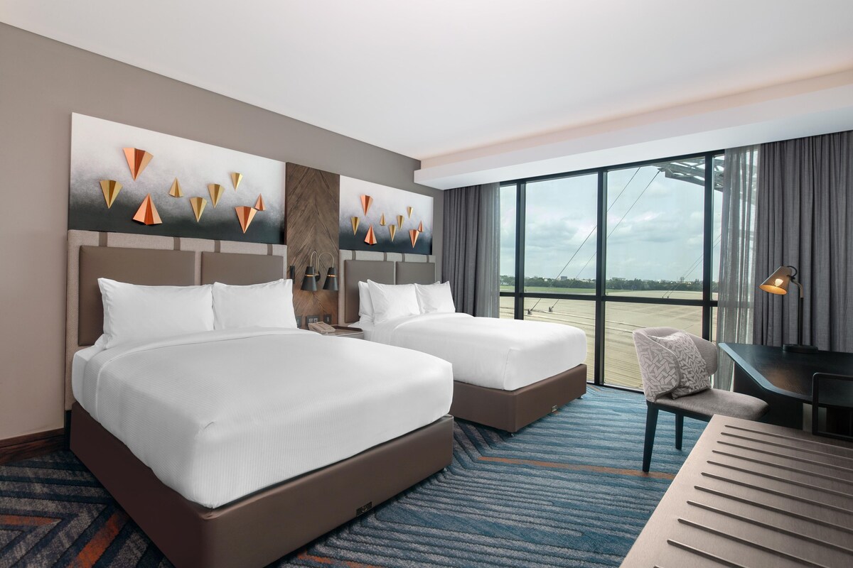 A hotel room with two beds, a desk, a chair, and large windows with a view.