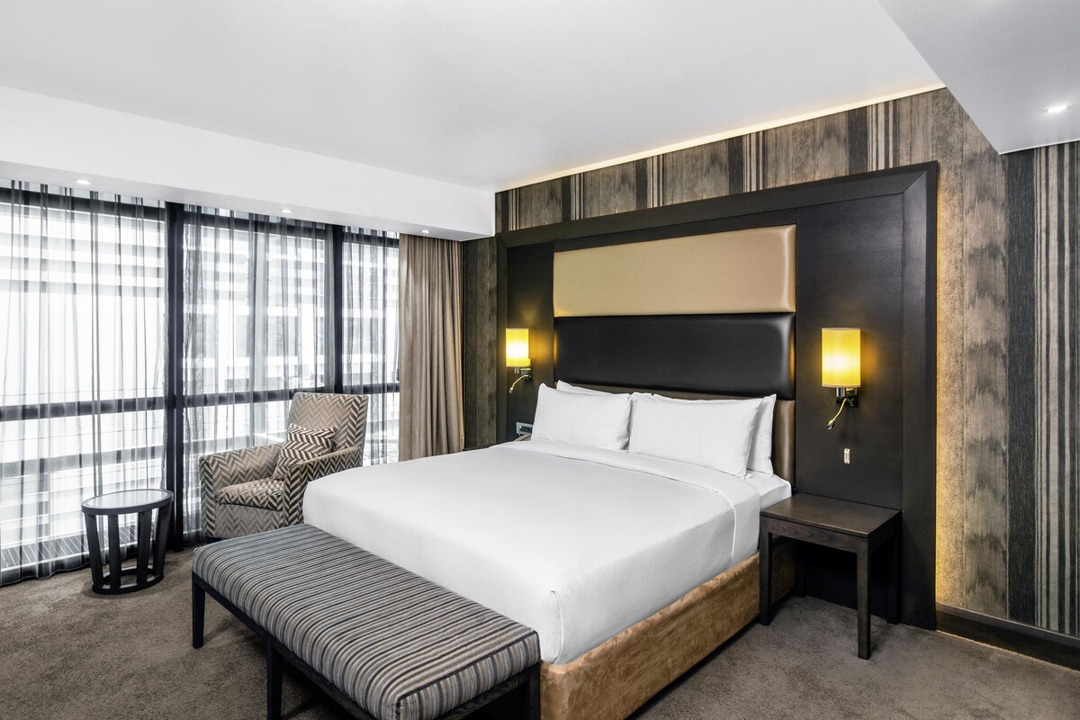 A hotel room with a large bed, a bedside table, a chair, a small table, and a bench.