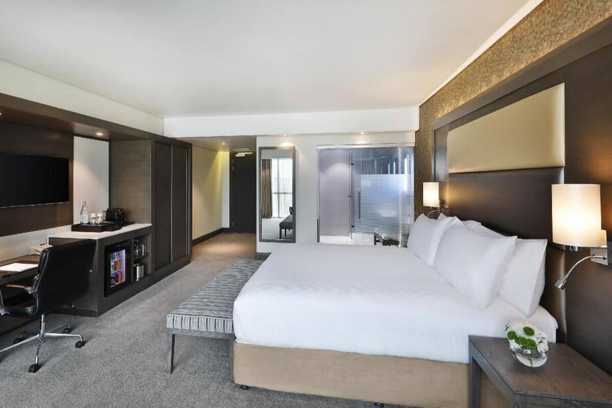 Habitación de hotel con una cama grande, un escritorio con silla, televisor y un baño visible a través de una puerta abierta.