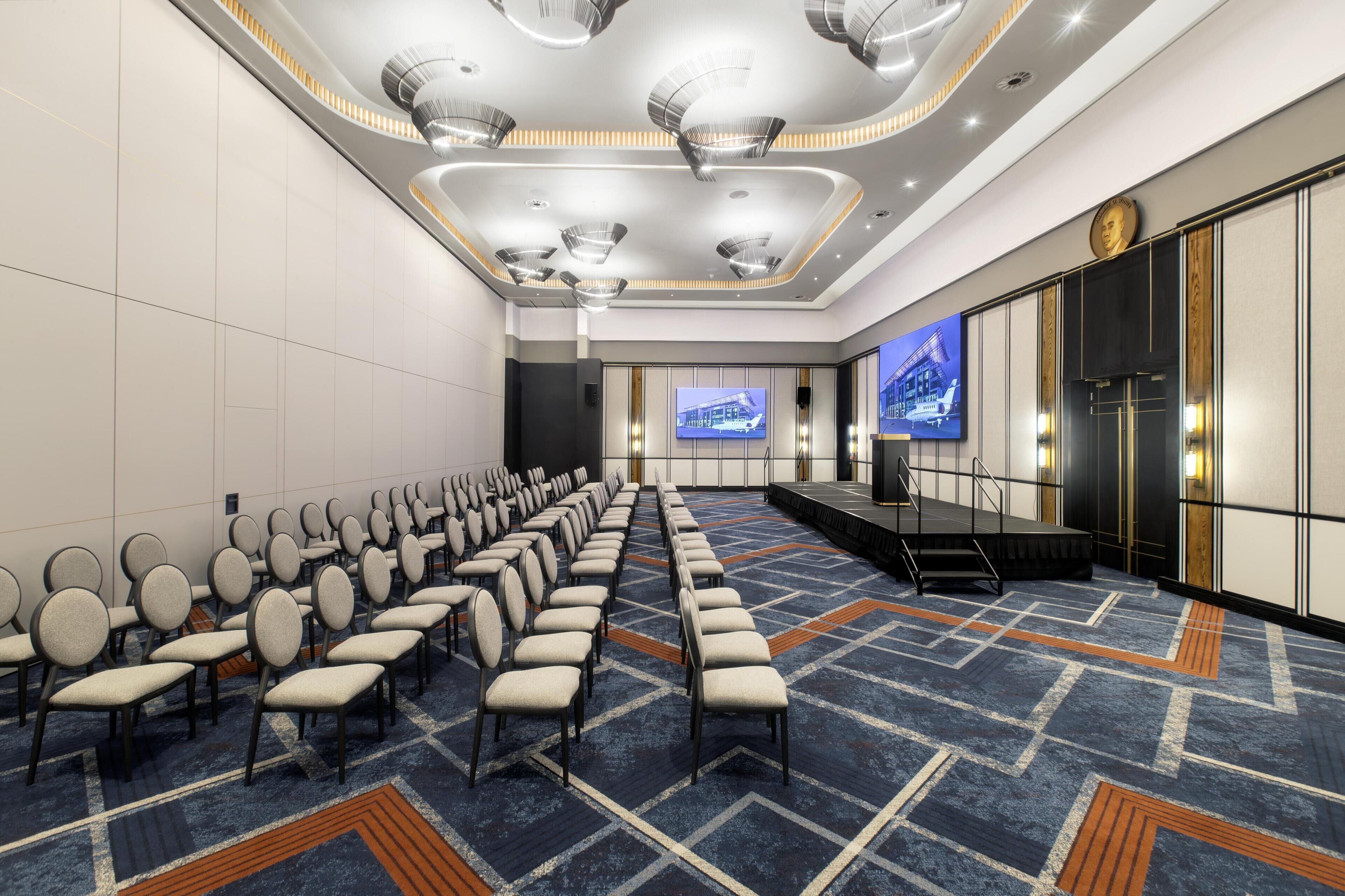 A conference room with rows of chairs, a stage, and a large screen.