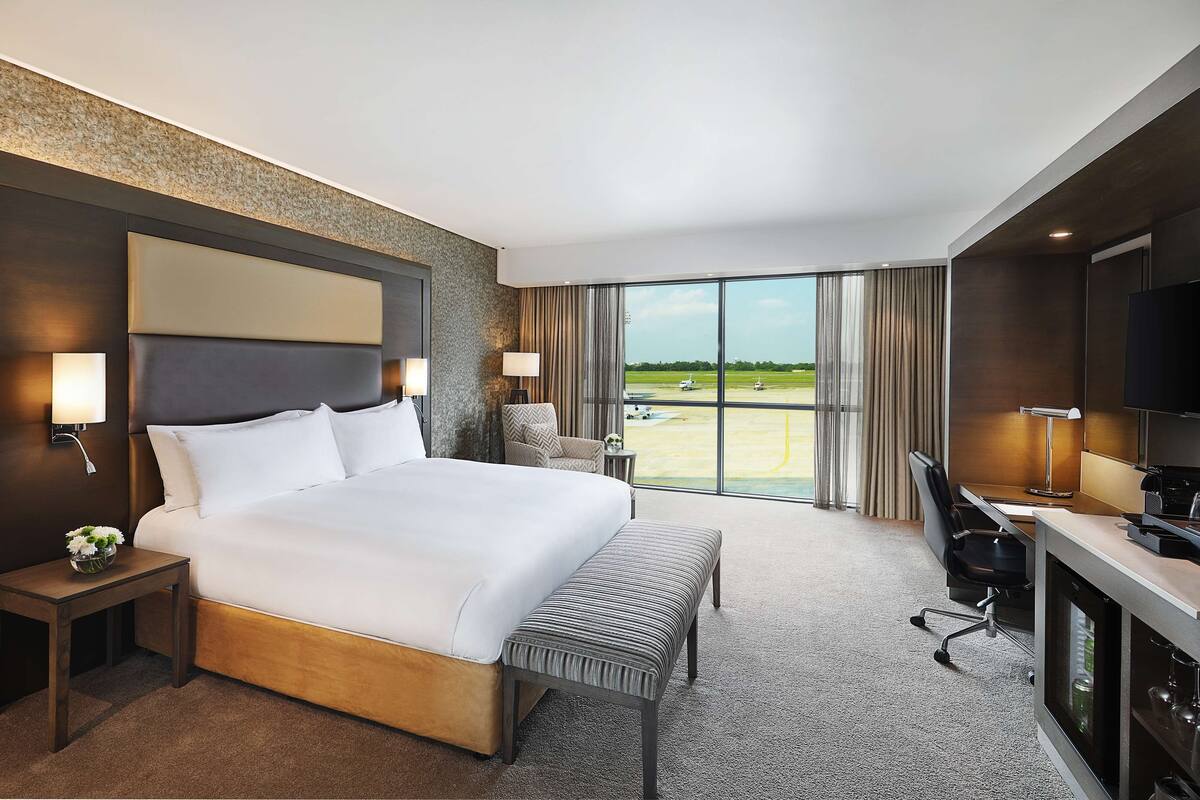 Habitación de hotel con una cama grande, un escritorio, una silla, un televisor y vistas al aeropuerto.
