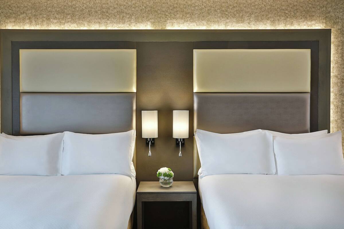 Una habitación de hotel con dos camas, una mesita de noche con un jarrón de flores e iluminación en la pared.