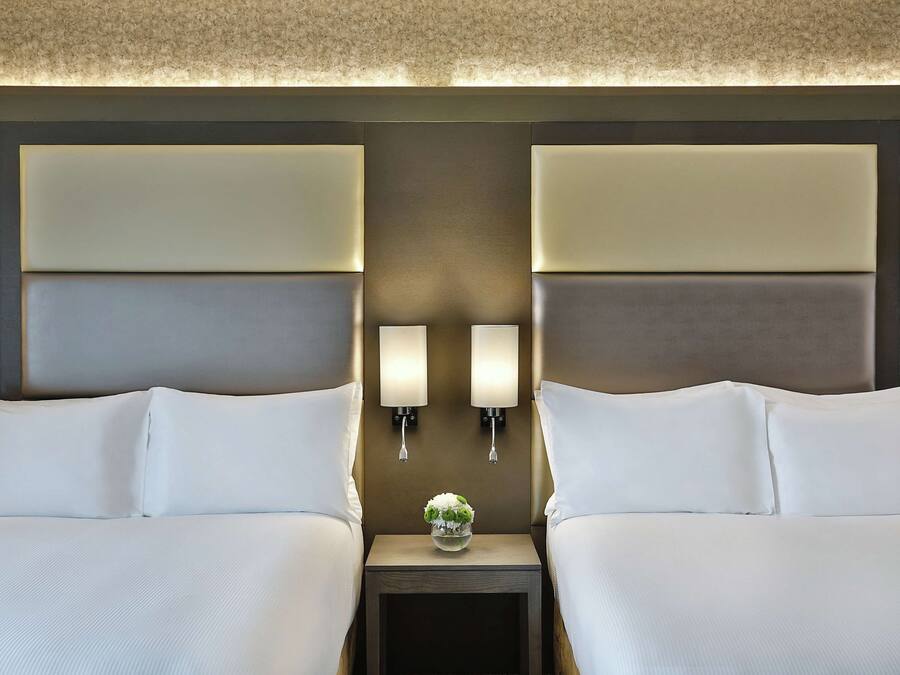 A hotel room with two beds, a nightstand with a vase of flowers, and wall-mounted lights.