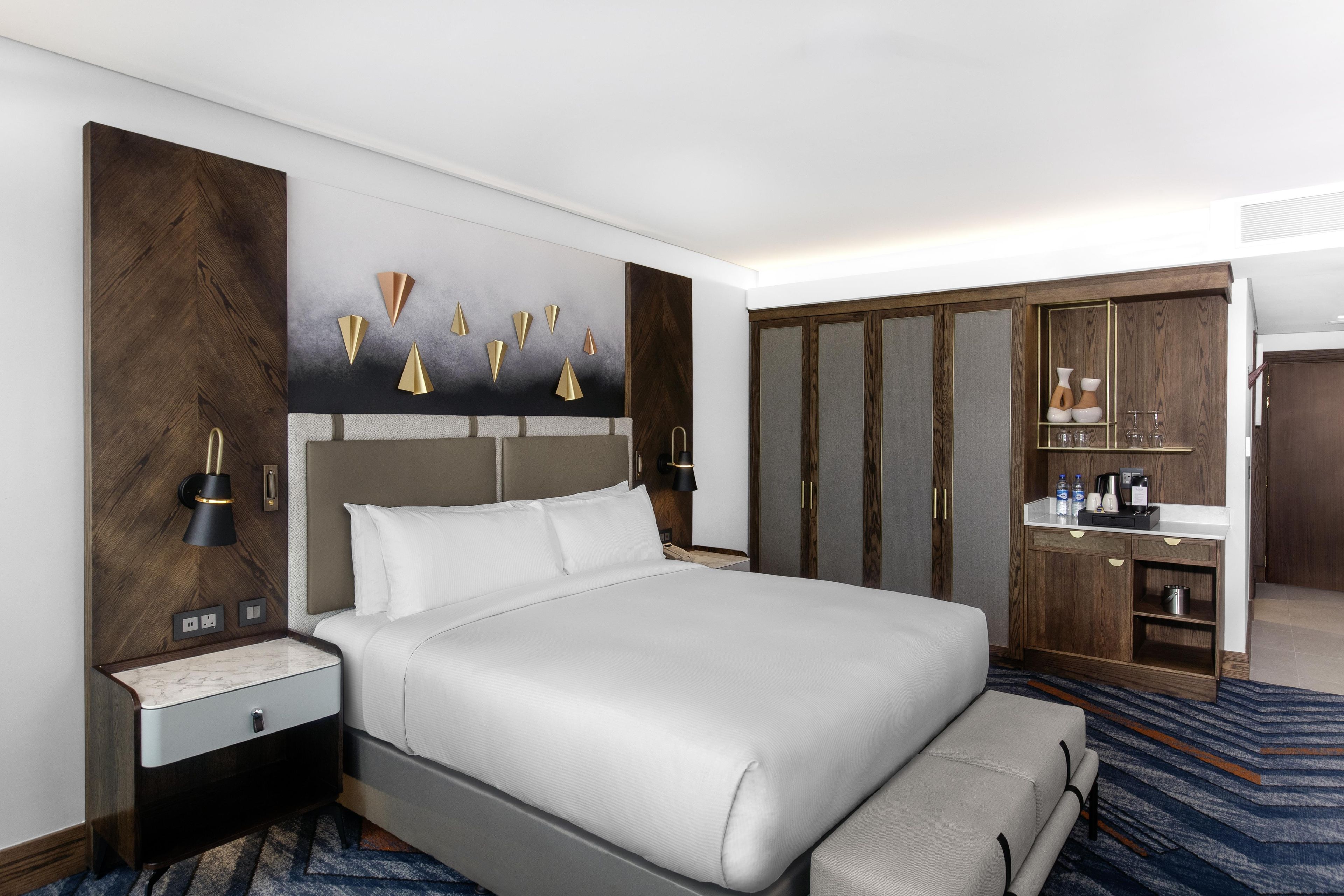 A modern hotel room with a large bed, a nightstand, a dresser, and a vanity with a mirror.