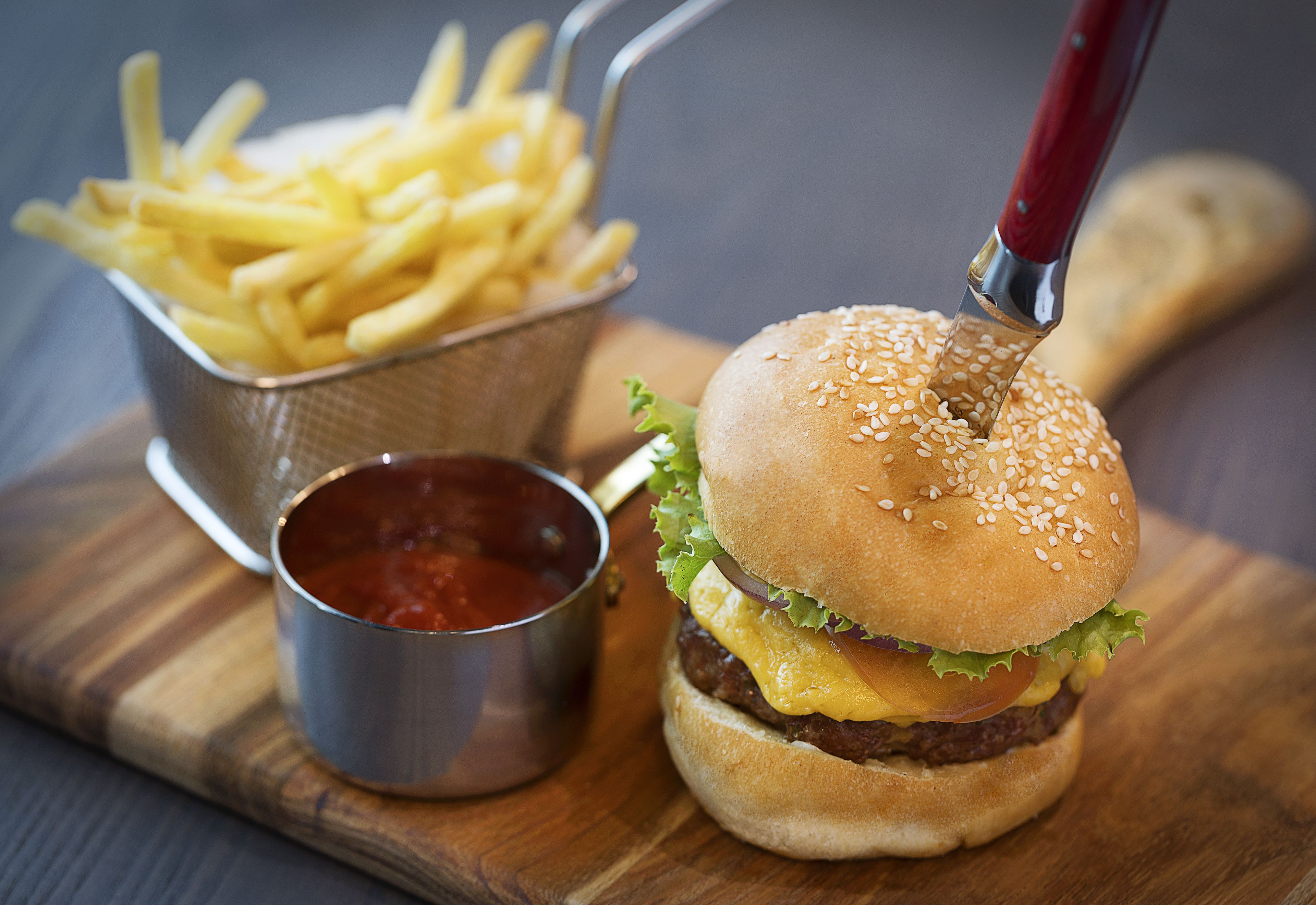 Een hamburger met sesamzaadbolletje, kaas, sla en een rundvleespatty, vergezeld van een klein metalen bekertje ketchup en een mandje friet.