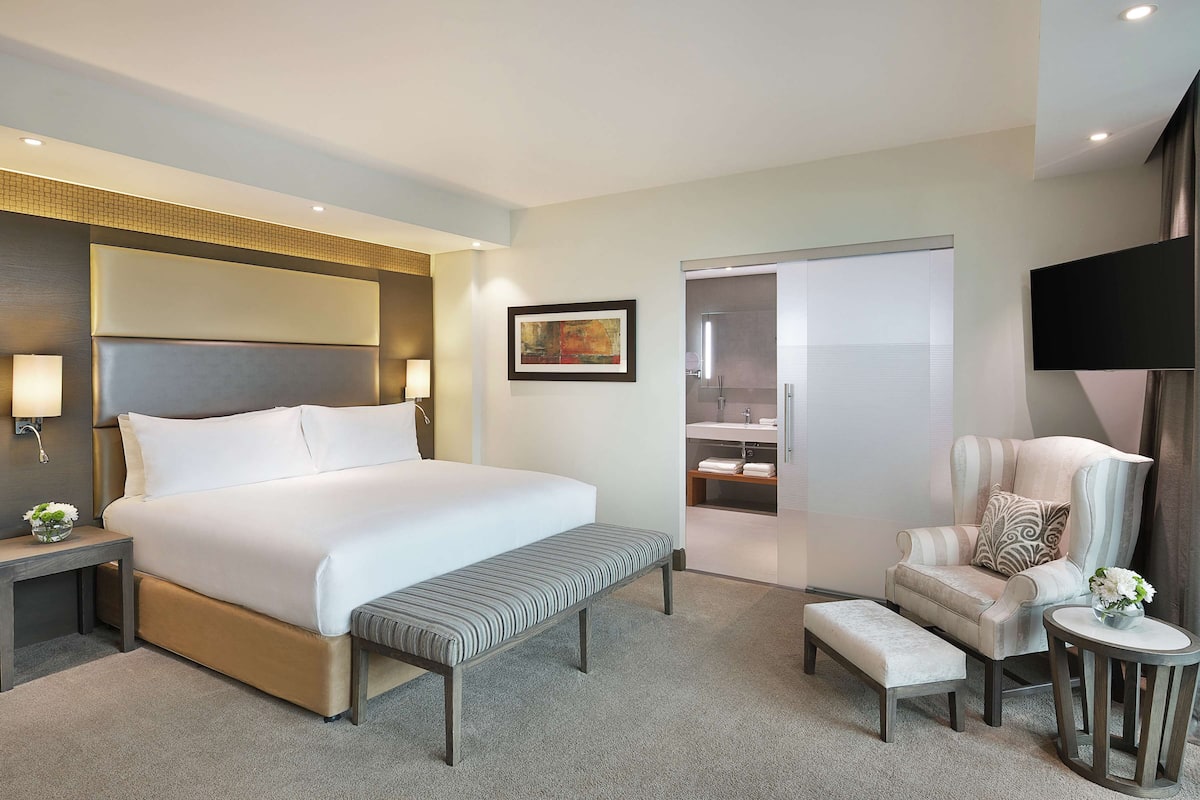 Habitación de hotel con una cama grande, mesitas de noche, un banco, una silla, una pequeña mesa con un florero y un televisor en la pared.