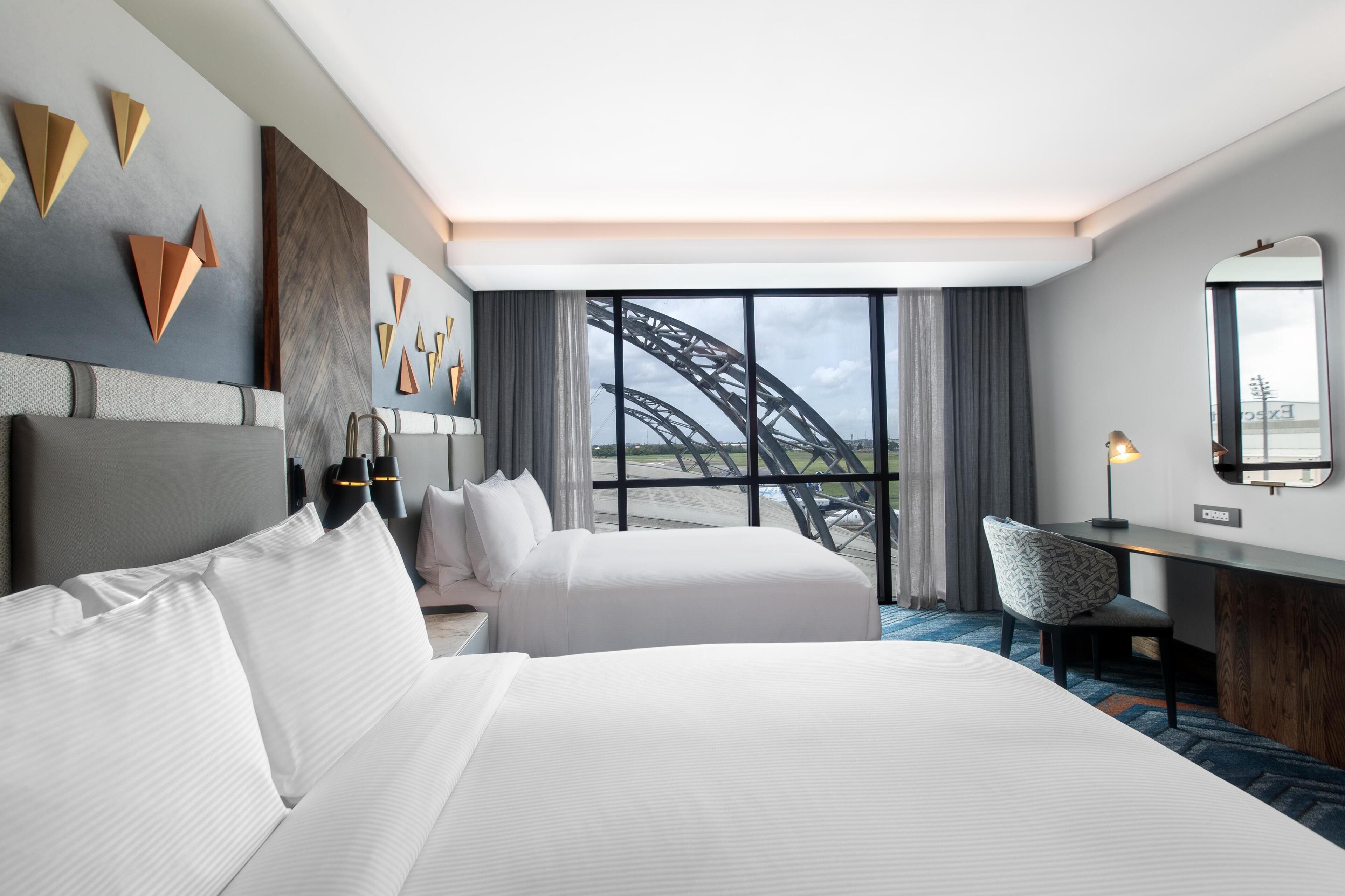 A hotel room with two beds, a desk, a chair, a large window with a view of a bridge, and wall art.