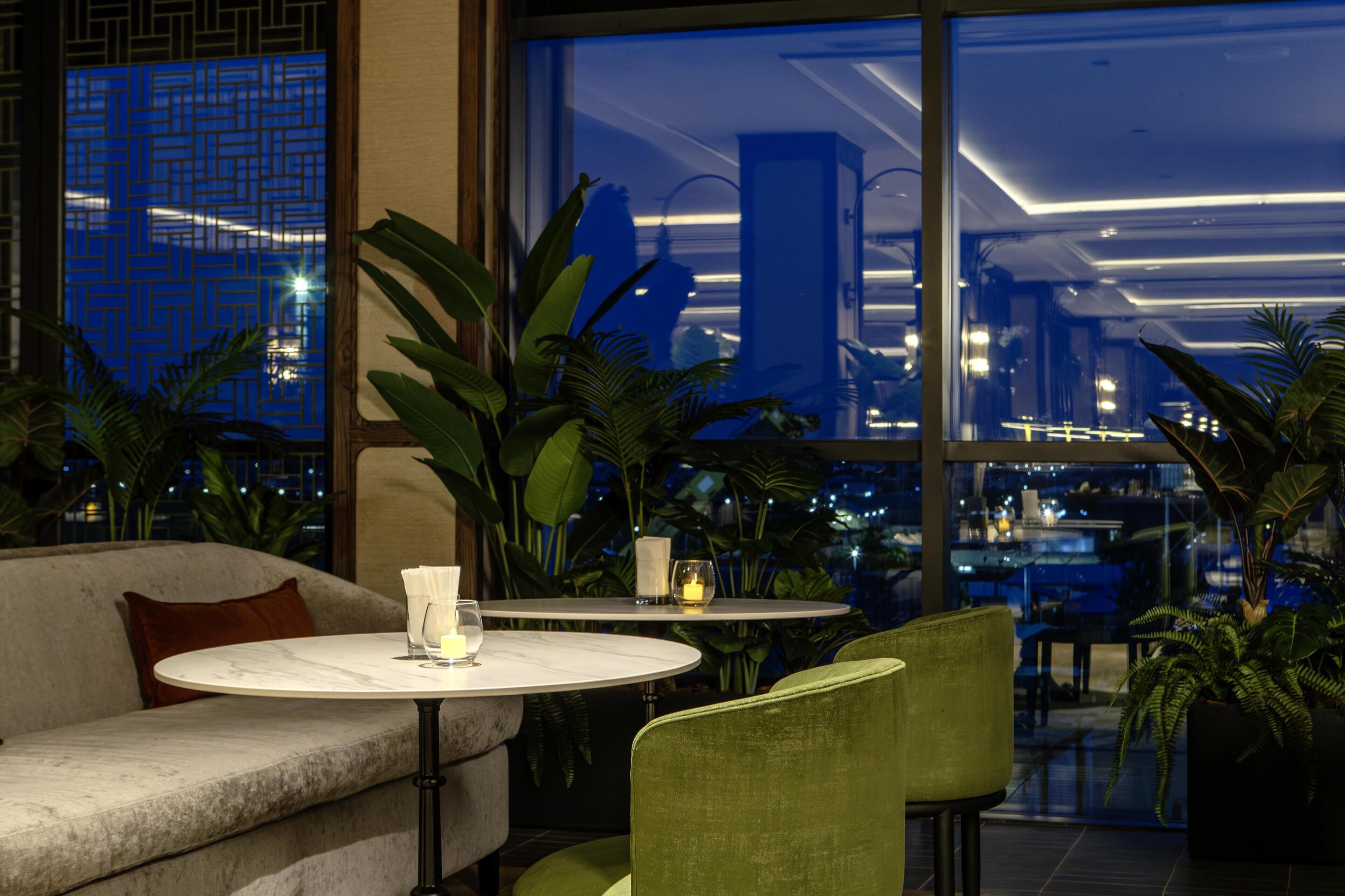 Un intérieur moderne avec un canapé, une table ronde et des chaises, de grandes fenêtres donnant sur la ville et des plantes d’intérieur.