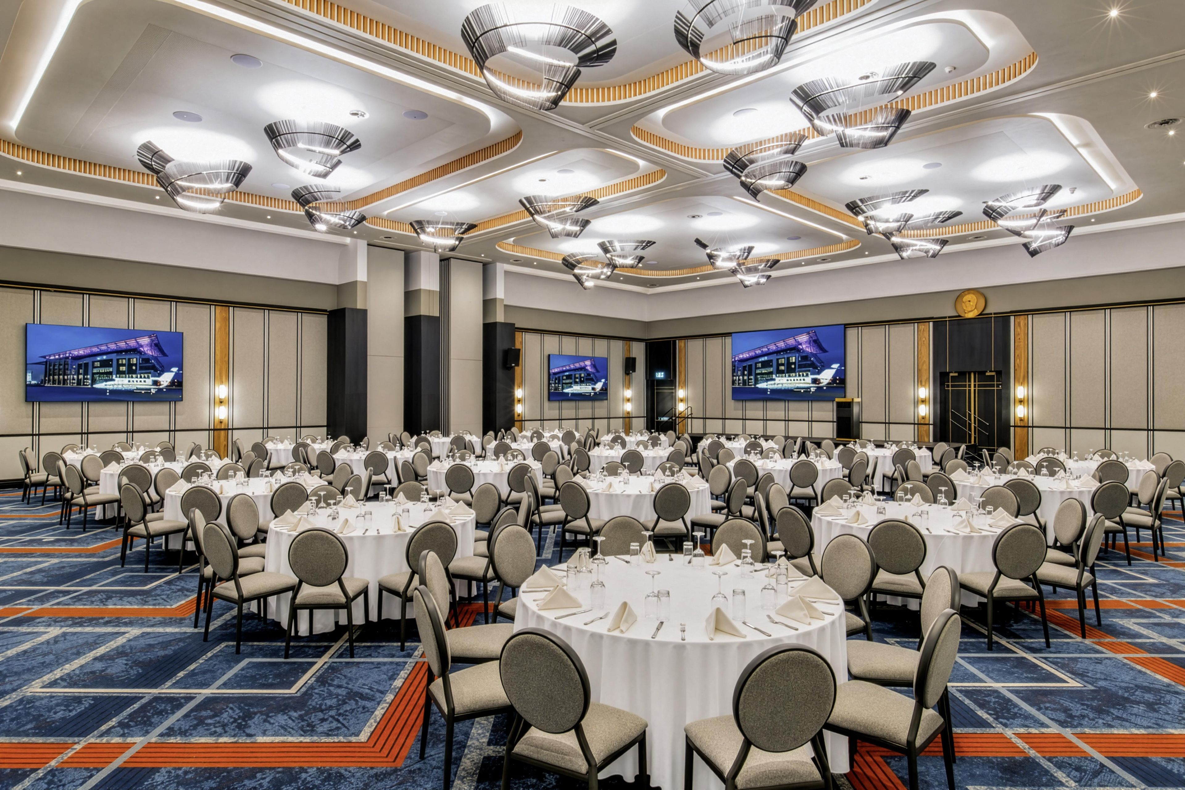 A large conference room with round tables, chairs, and multiple screens on the walls.