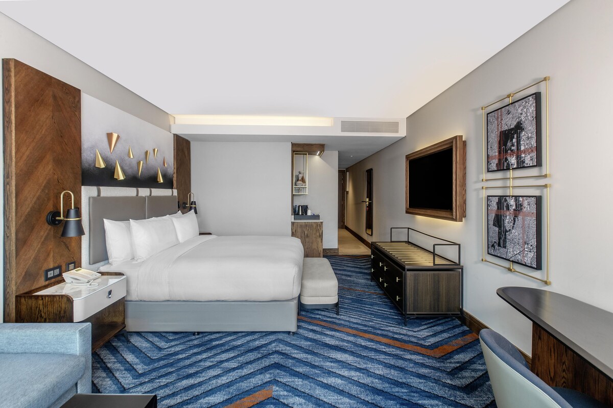 A hotel room with a bed, a desk, a chair, a TV, and artwork on the walls.