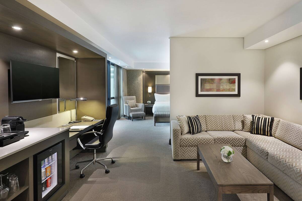 Habitación de hotel con televisión de pantalla plana, escritorio con lámpara, sofá, mesa de centro y un cuadro en la pared.