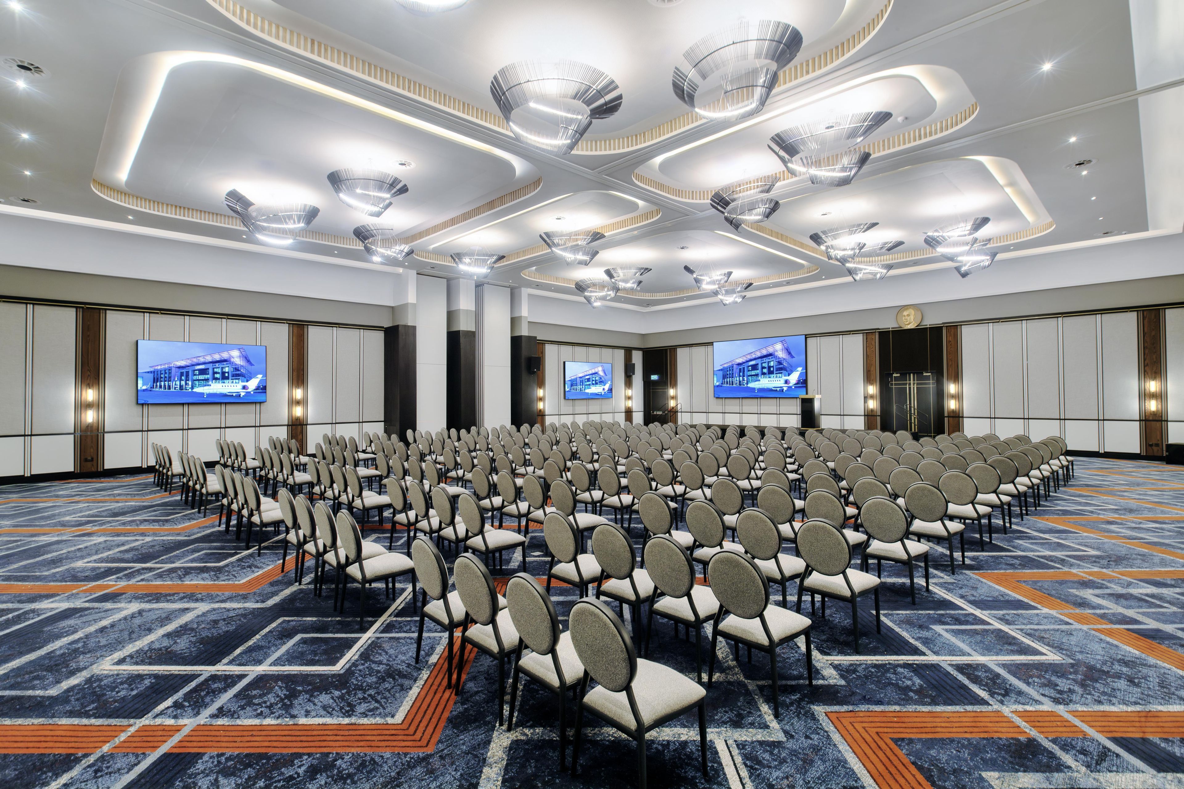 Un salón de conferencias con filas de sillas, pantallas grandes e iluminación de techo moderna.