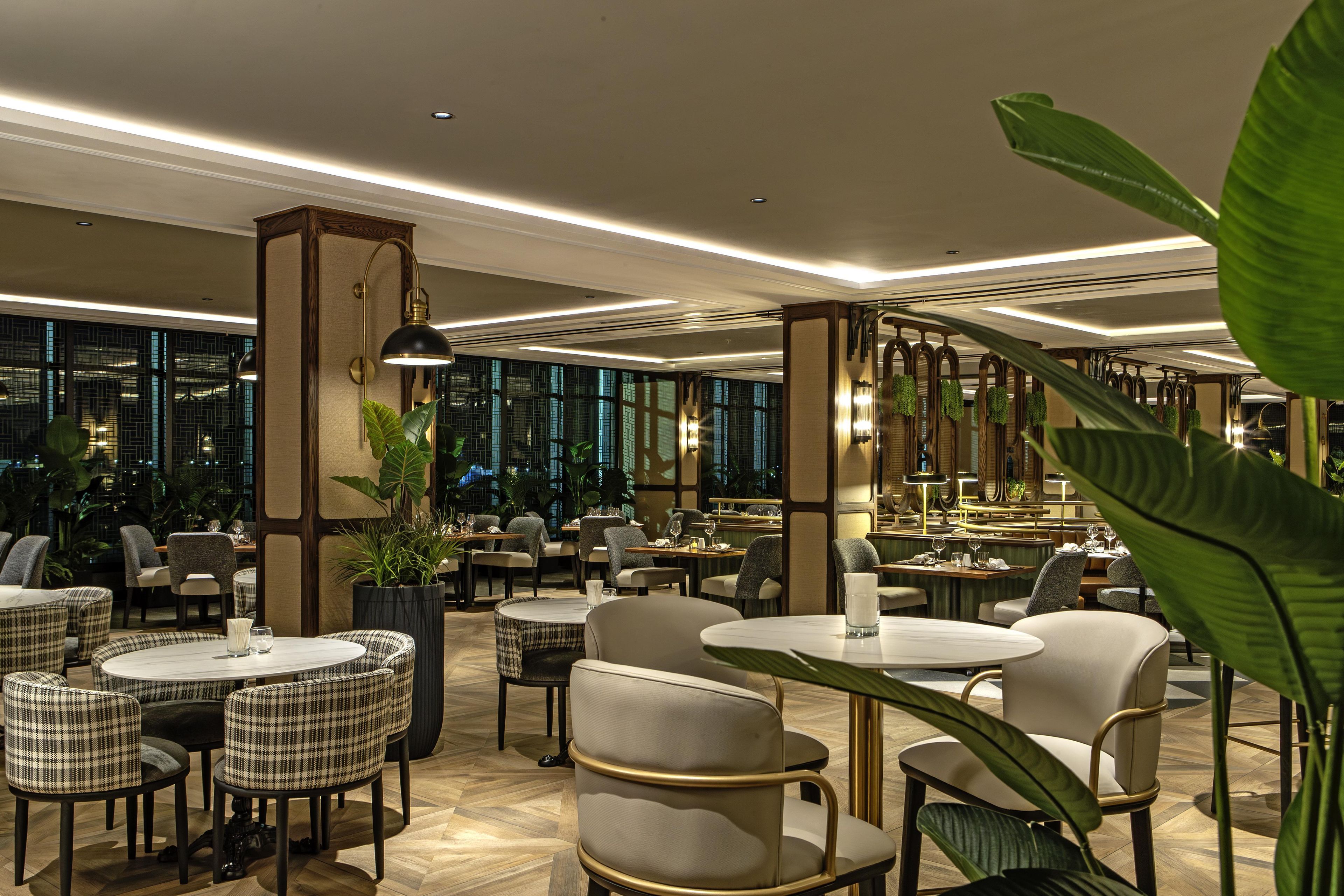 Ein modernes Restaurant mit runden Tischen und Stühlen, großen Fenstern und Zimmerpflanzen.