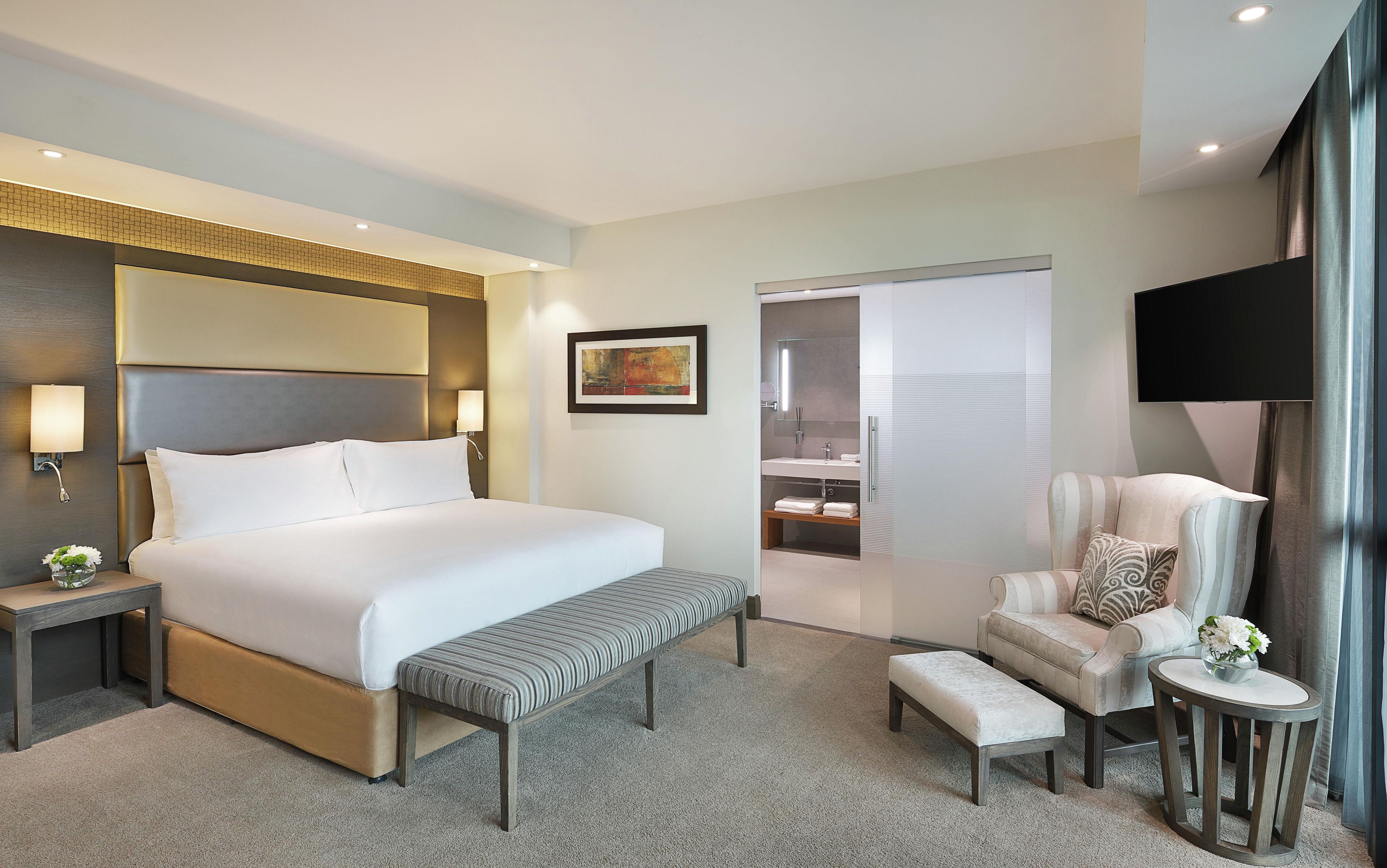A hotel room with a large bed, a bench, a chair, a small table with a vase, a painting on the wall, and a television.
