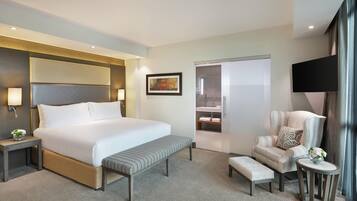 A hotel room with a large bed, a bench, a chair, a small table with a vase, a painting on the wall, and a television.