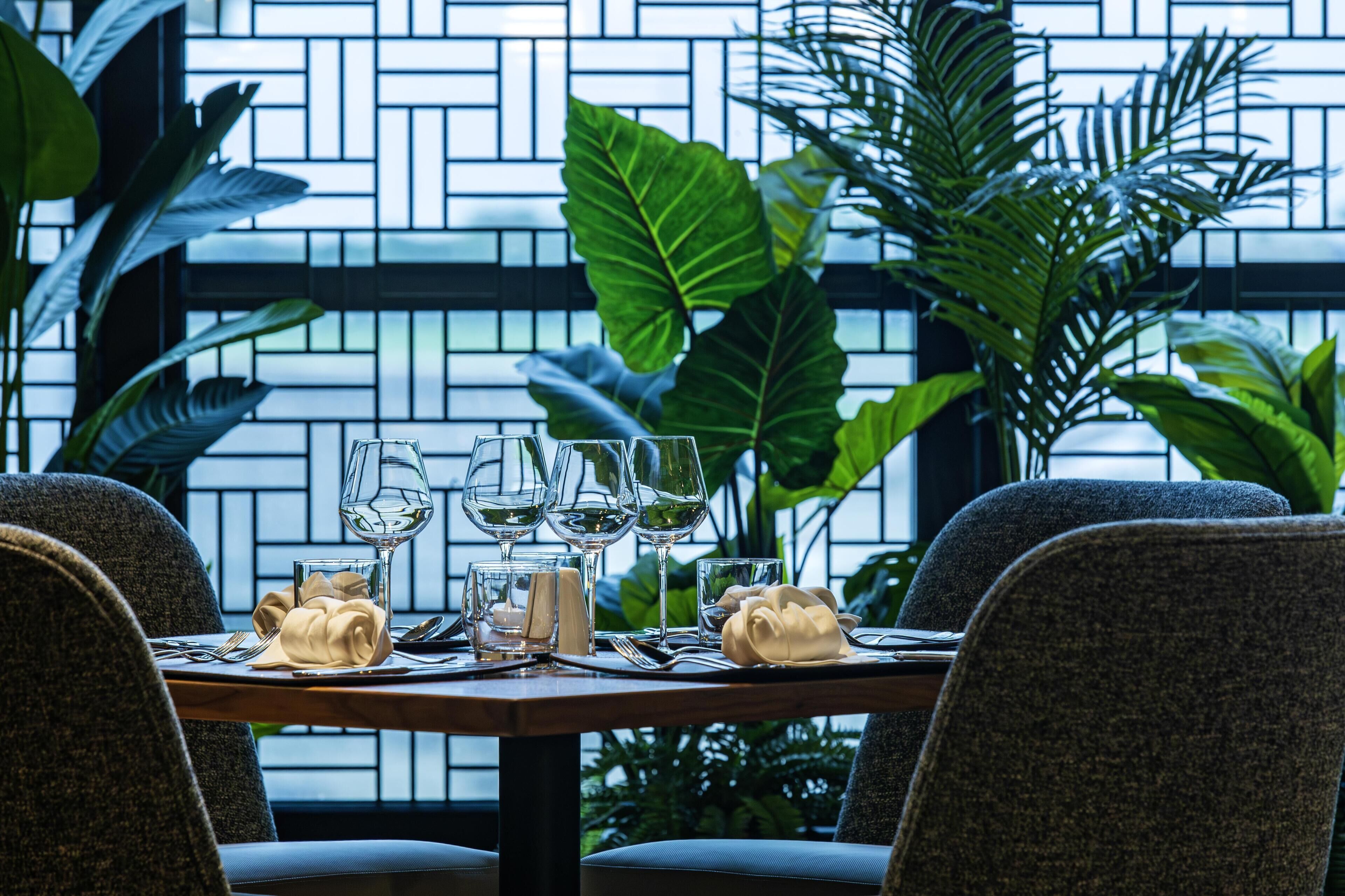 A dining table set with wine glasses, plates, and napkins, surrounded by chairs, with a view of a brick wall and greenery.
