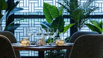 A dining table set with wine glasses, plates, and napkins, surrounded by chairs, with a view of a brick wall and greenery.