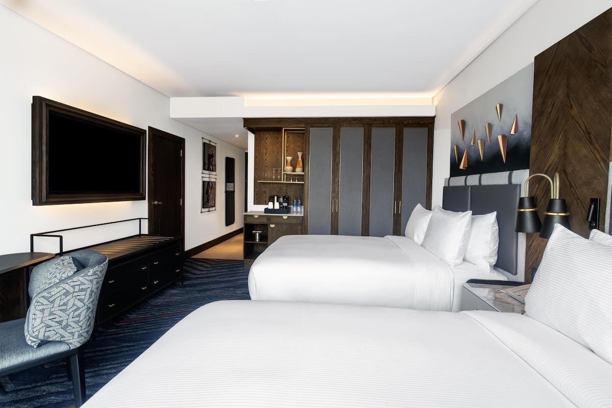 Habitación de hotel con dos camas, un escritorio, una silla, un televisor, un minibar y un cuadro en la pared.