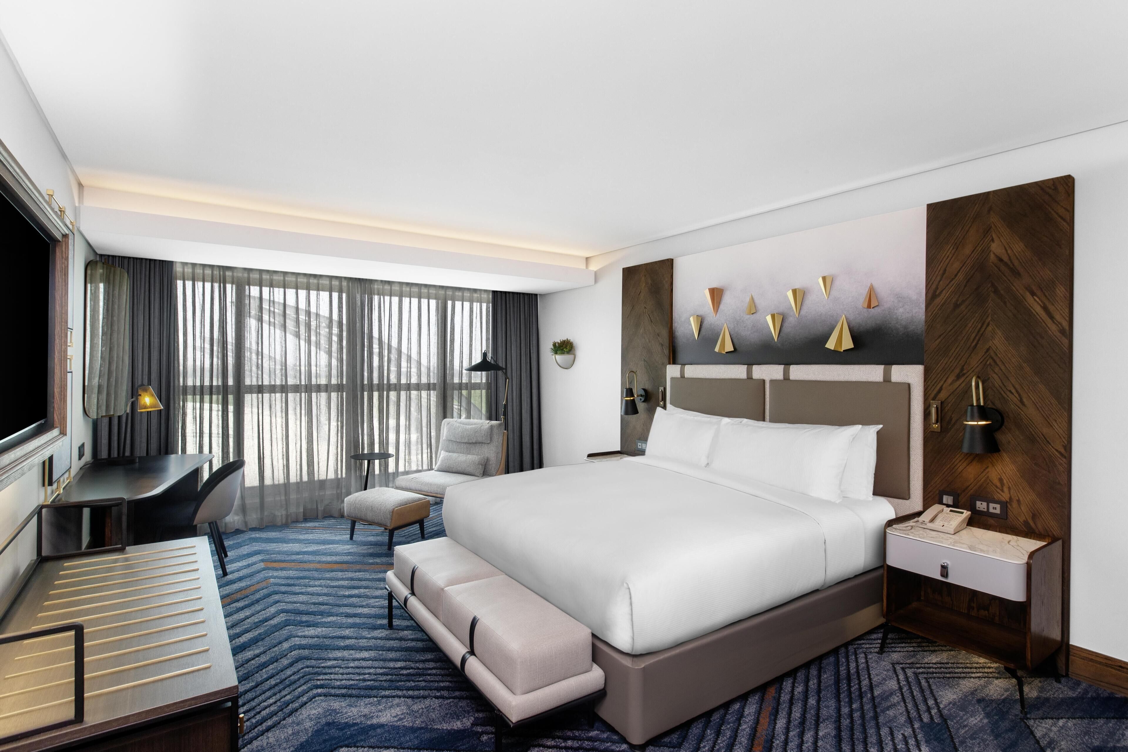 A modern hotel room with a large bed, a desk, a chair, and a TV.