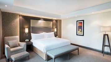 Quarto de hotel com cama, cadeira, banqueta, mesa de cabeceira com abajur e um quadro na parede.