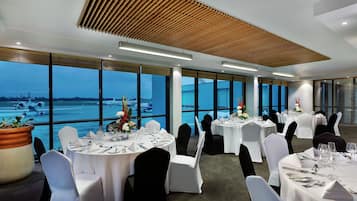 A conference room with round tables, white tablecloths, and black chairs. Large windows offer a view of an airport.
