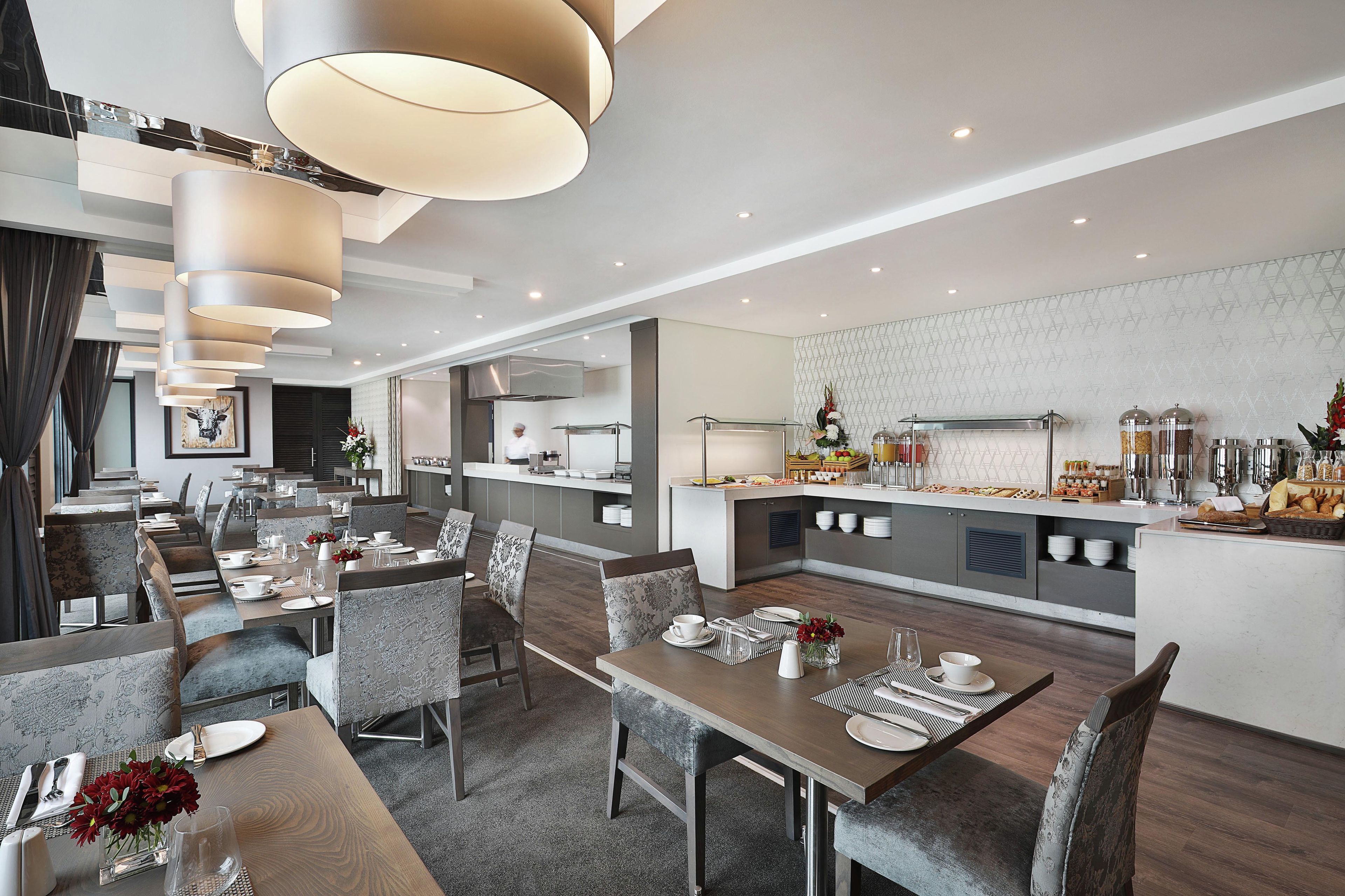 Et moderne hotelspiseområde med flere borde, stole og et centralt køkkenområde.