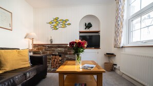 Ein Wohnzimmer mit Kamin aus Stein, einem Holztisch mit Blumenvase, einer schwarzen Ledercouch und einem Fernseher an einer weißen Wand.