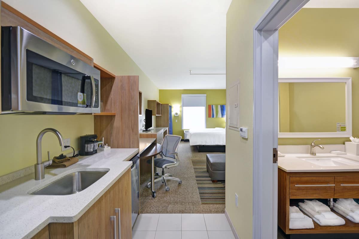 Une chambre d’hôtel moderne dotée d’une kitchenette, d’un grand lit, d’une salle de bain avec un lavabo et un miroir, et d’un petit espace bureau.