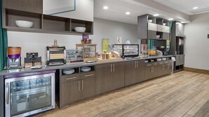 A self-service breakfast area with a refrigerated display case, a waffle maker, and a variety of food items. - Home2 Suites by Hilton Dallas DeSoto (DeSoto)
