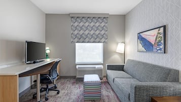 A hotel room with a desk, chair, TV, couch, and a window with patterned curtains.