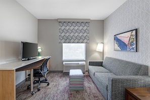 A hotel room with a desk, chair, TV, couch, and a window with patterned curtains. - Home2 Suites by Hilton Dallas DeSoto (DeSoto)