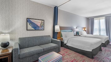 A hotel room with a sofa, a bed, a colorful ottoman, a painting, and a patterned carpet.